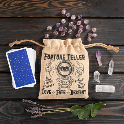 A foux burlap puch with drawstring closure on a wood background. Pouch has black graphics showing a crystal ball with the all-seeing eye, hands, and the words "Fortune Teller - Love - Fate - Destiny." Crystals, runes and a tarot deck surround the pouch.