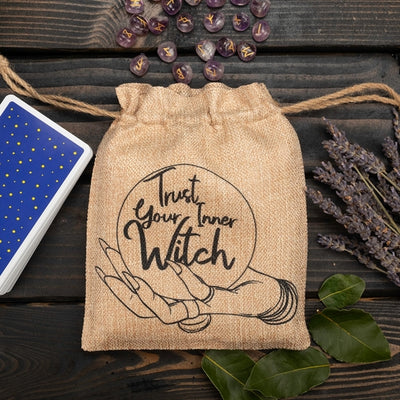Faux burlap pouch with drawstring closure and black graphics against a wood background. Pouch has an image of a hand holding a crystal ball printed with the words "trust your inner witch." The pouch is surrounded by runes, a deck of cards, and dried lavender. 
